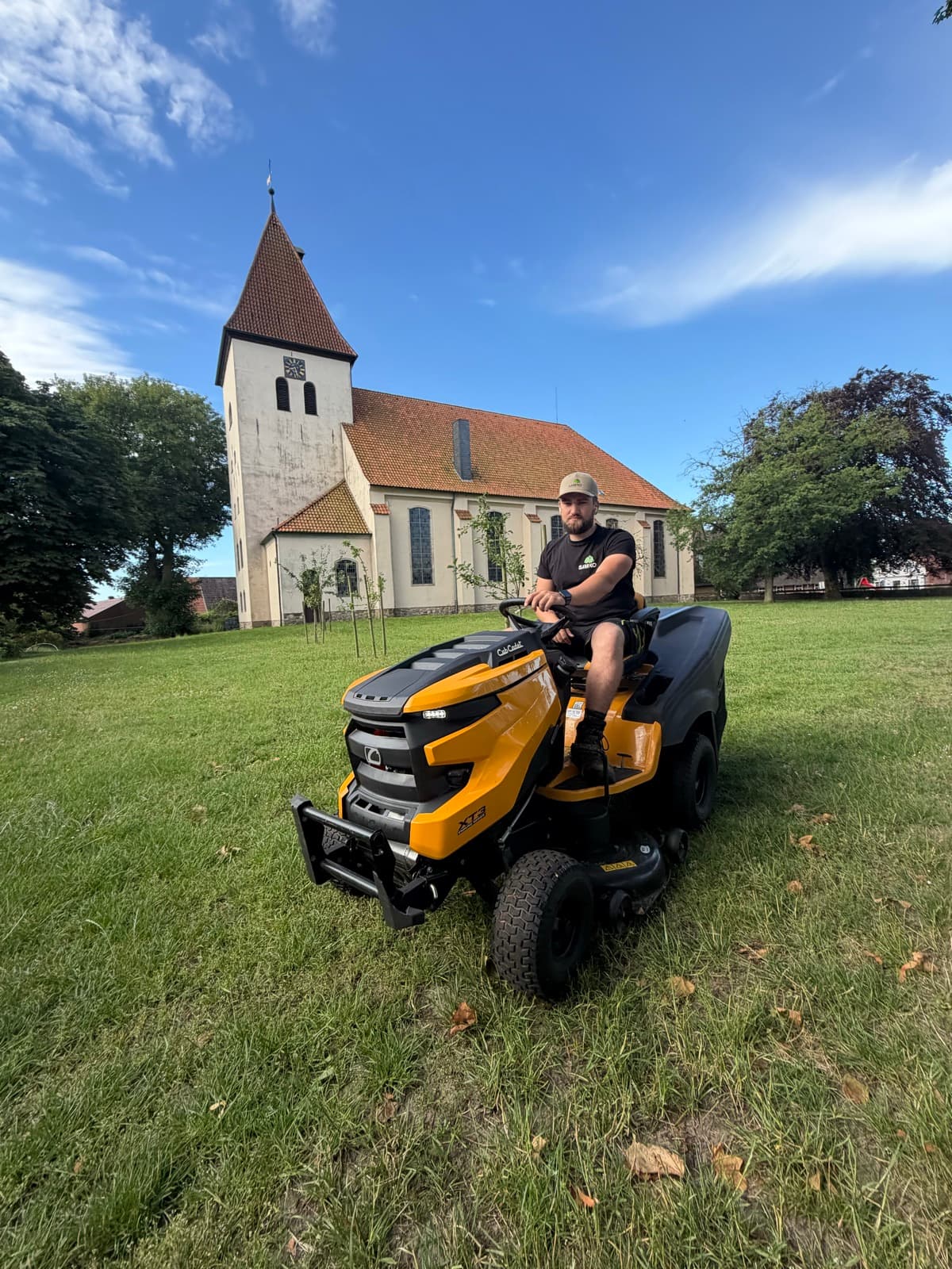 SAMKO-Mitarbeiter mäht mit dem Aufsitzmäher die Rasenfläche vor einer Dorfkirche