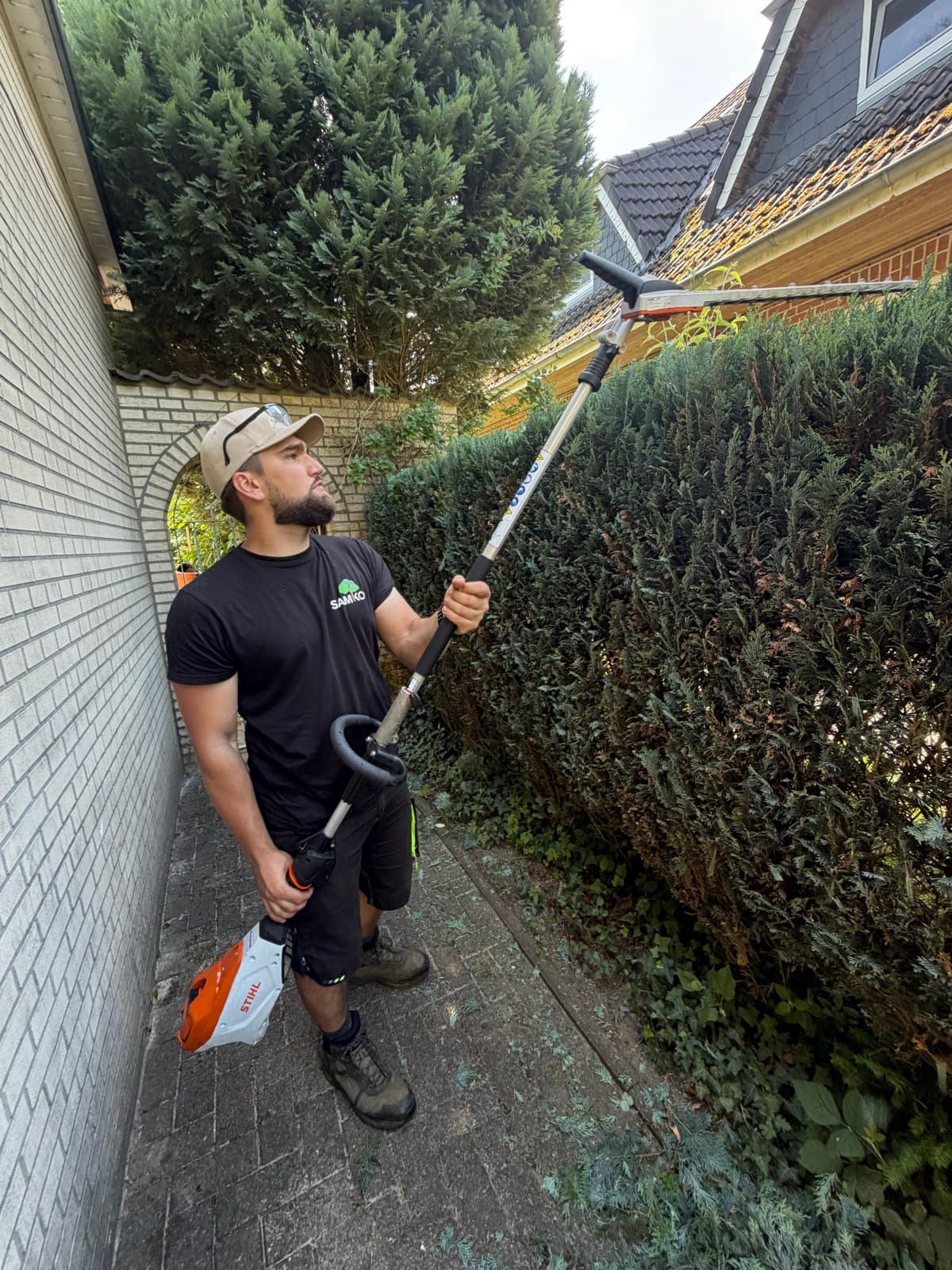 SAMKO-Mitarbeiter schneidet eine hohe Hecke neben einem Wohnhaus mit der Teleskop-Heckenschere